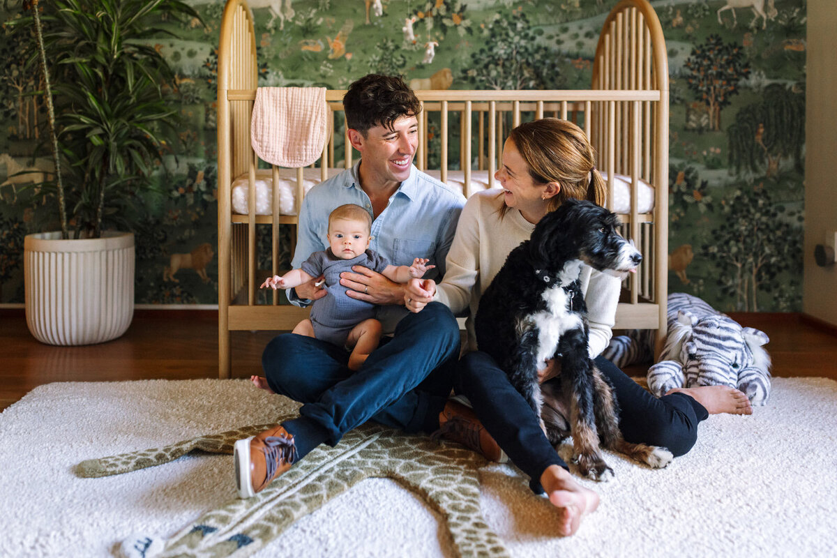 dad holding baby and mom holding dog sitting on floor in nursery in front of crib