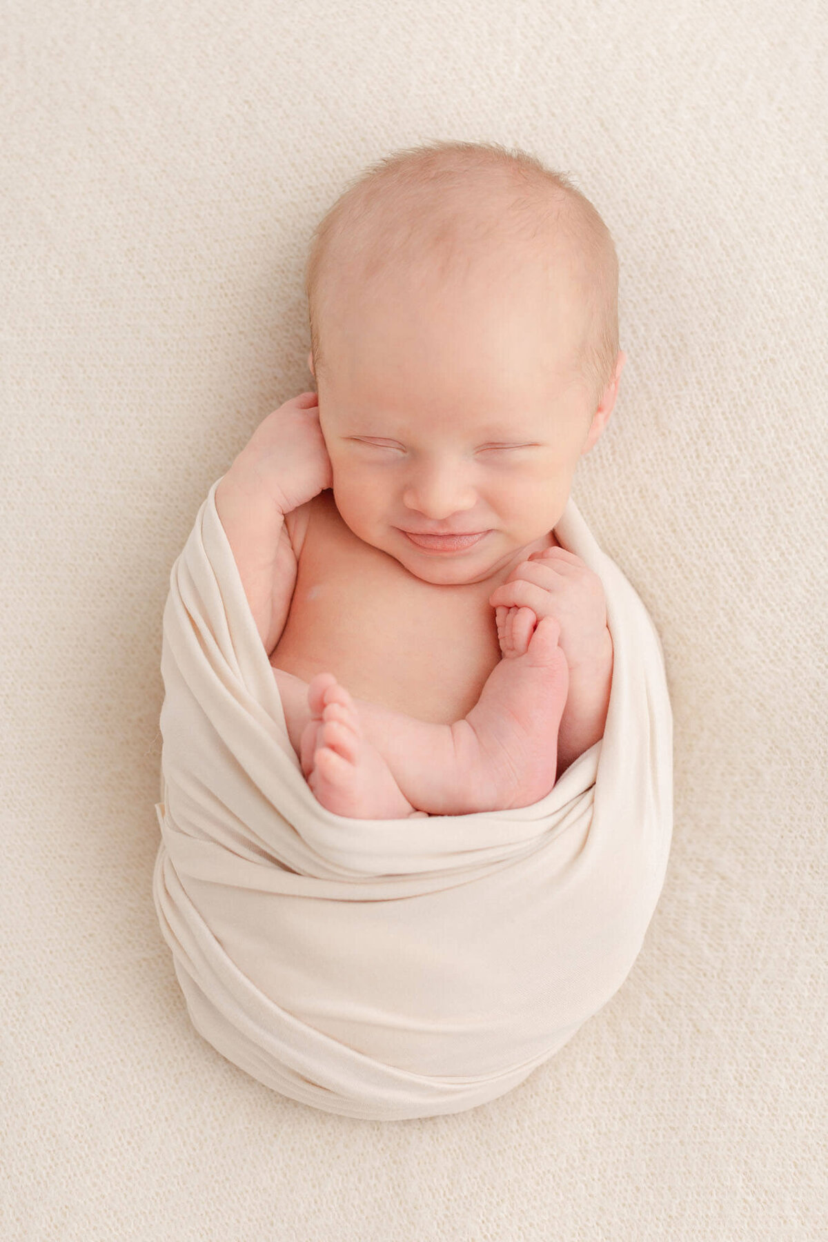 Newborn Babe laying on his back on a beige blanket with a beige wrap around his body. His arms and feet and tummy are showing and one hand is up by his cheek. He is sleeping and has a little grin on his face.