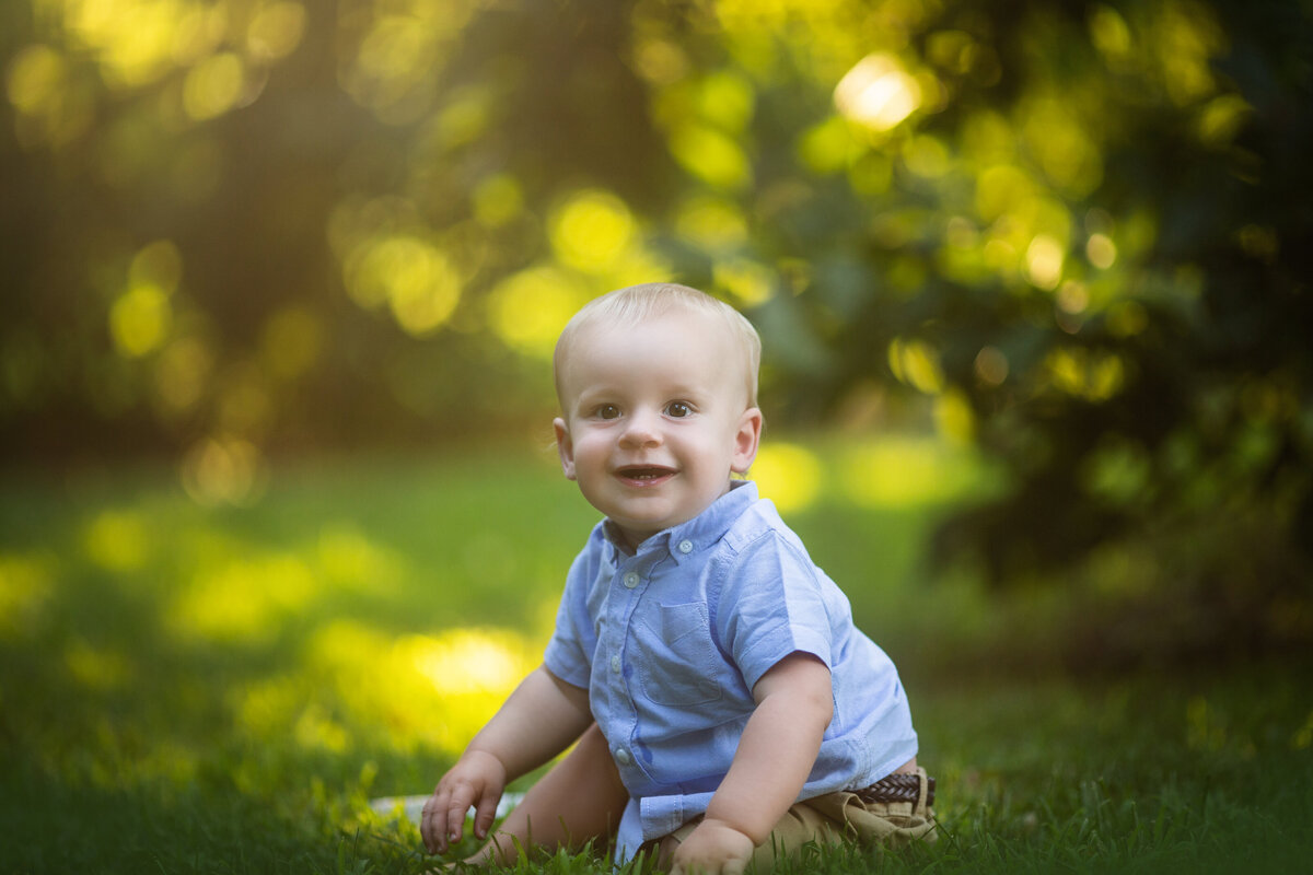 A young toddler boy sits in some grass playing in a blue shirt
