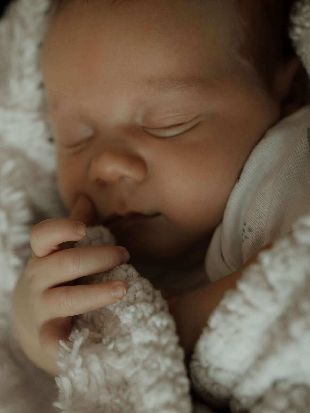 Baby sleeping soundly wrapped in a white blanket with his little hand resting near his mouth.