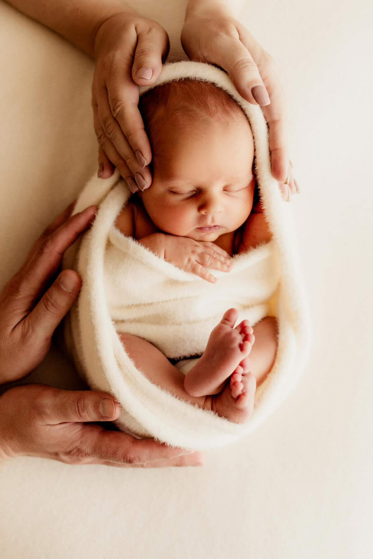 Mother and father place their hands on their newborn baby boy who is swaddled and wrapped in a cream fuzzy wrap, Edmond Oklahoma.
