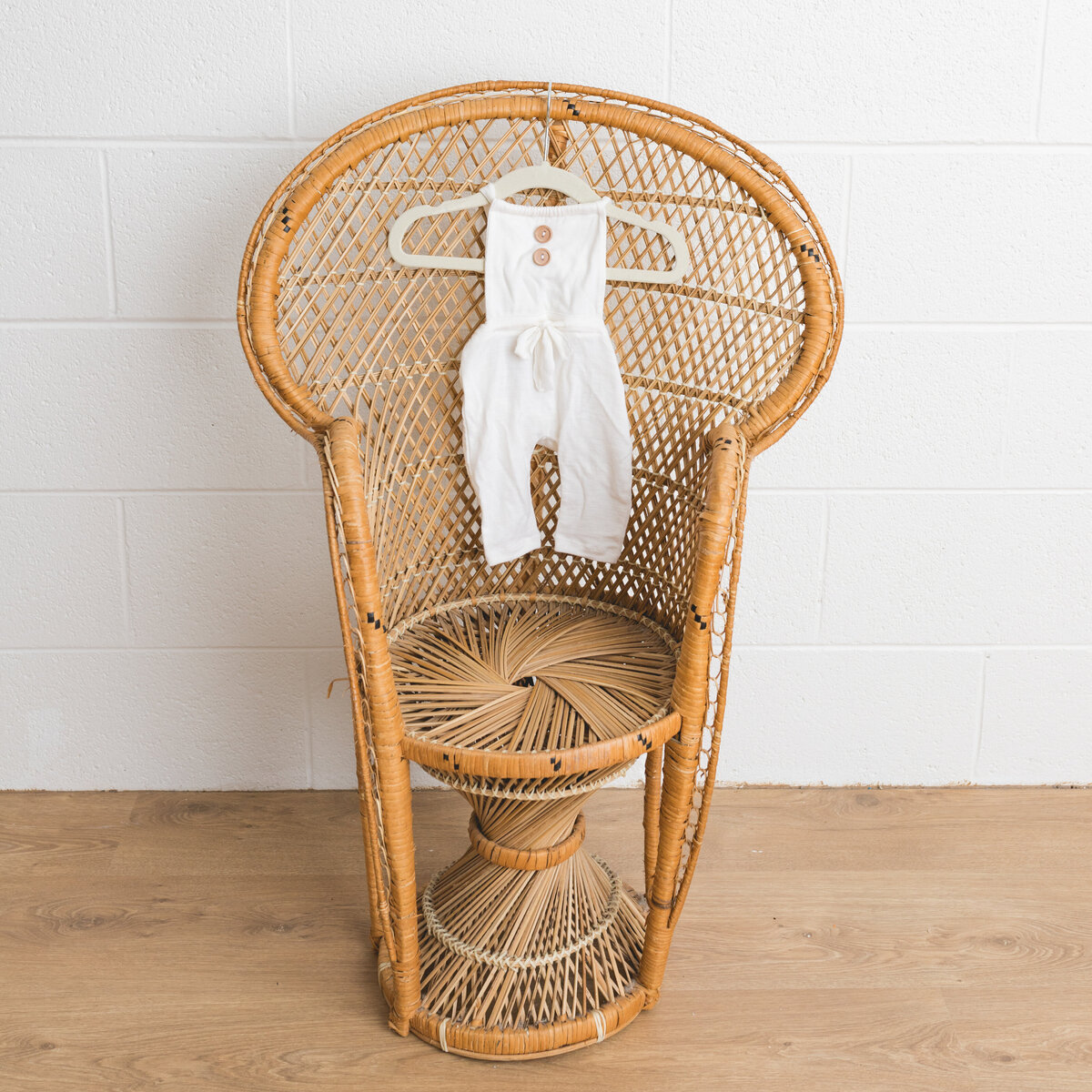 Image depicts a miniature peacock chair in a photography studio on the chair hangs a tiny newborn overalls handmade in white cotton with a halter style tie and brown wooden buttons.  This image is a sample of Lauren Vanier Photography's newborn client wardrobe. Image taken in Hobart Tasmania