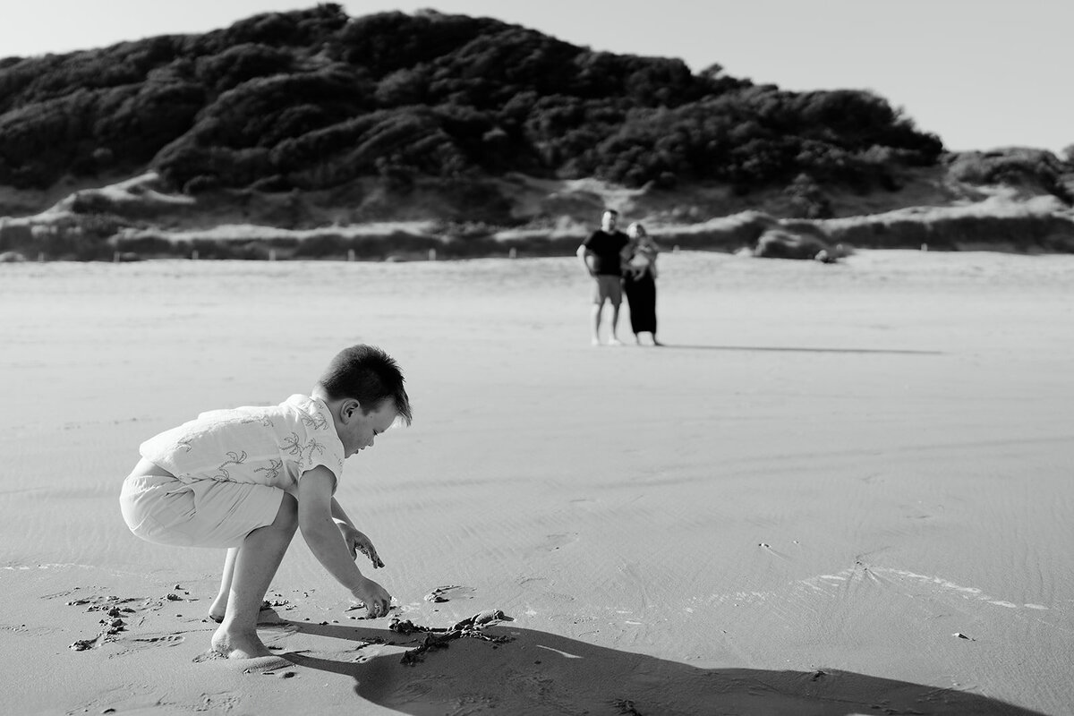 Black and white beach family photo in Geelong featuring a young boy playing in the sand with parents watching in the background near coastal dunes.