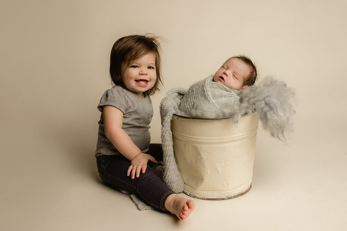 big sister sitting next to a bucket that holds her new baby brother