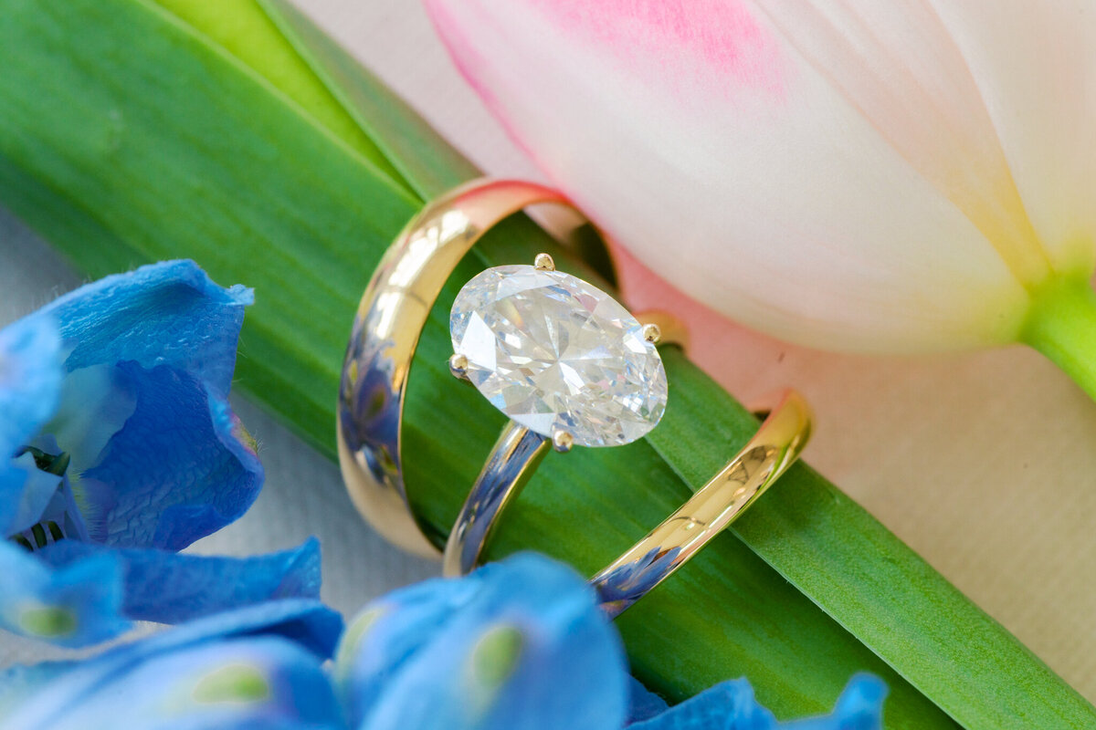 Close-up of oval diamond engagement ring and gold wedding band styled with a pink tulip and blue delphinium flowers.