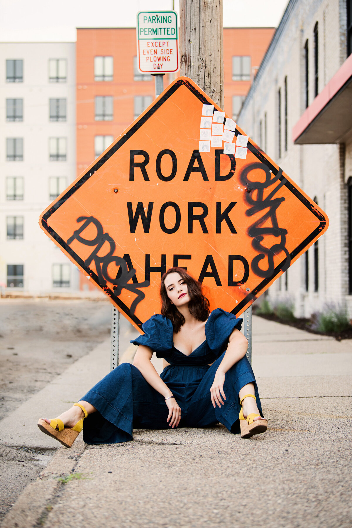 senior photo of girl in blue jumpsuit leaning against an orange road work sign in minneapolis