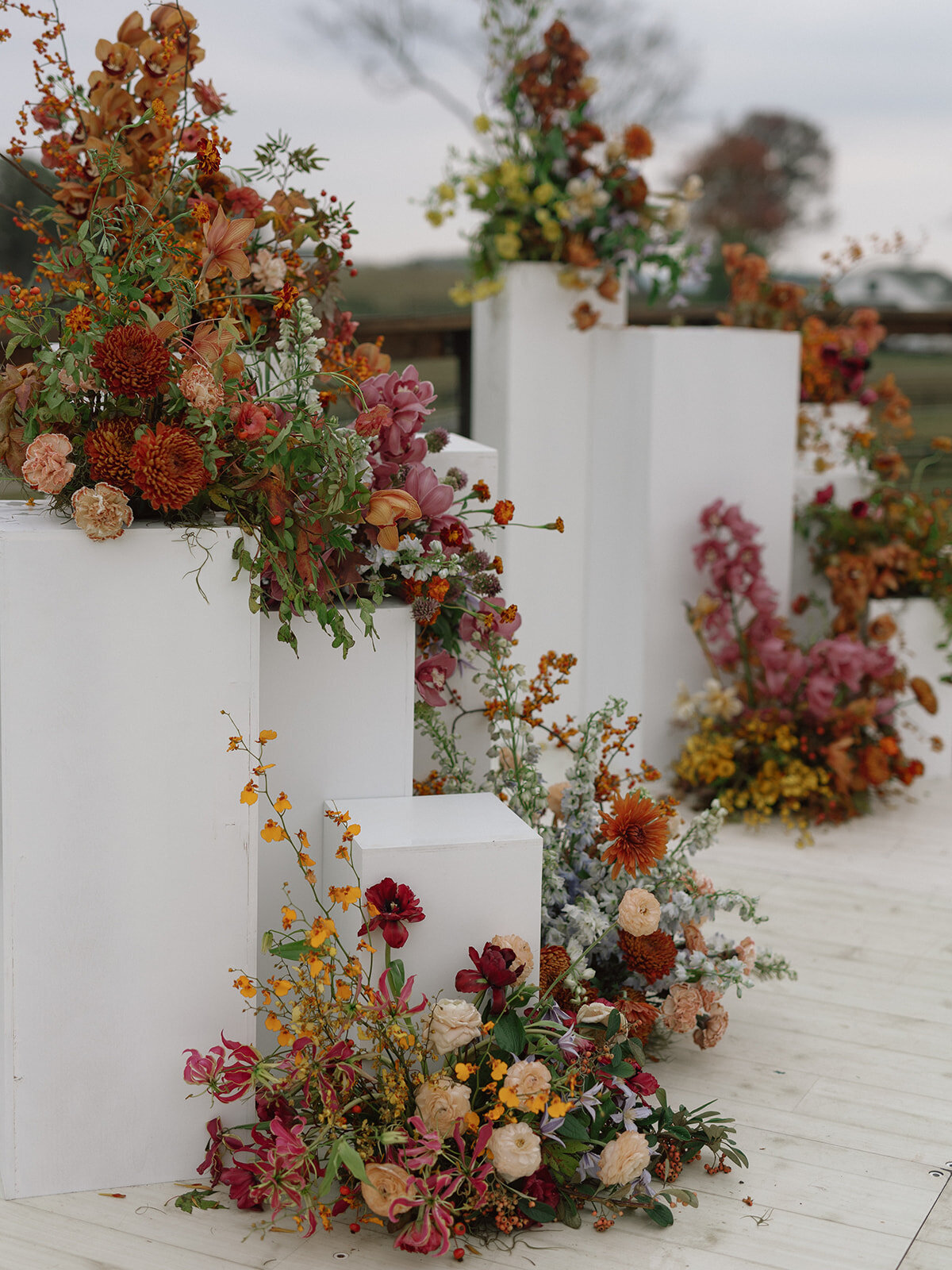 Sculptural columns wedding ceremony backdrop. Stair step white ceremony columns adorned with sculptural floral meadows in fall hues. Clear top tent build out for fall wedding reception in Tennessee hills. Wire threaded orchids, bittersweet berries, butterfly ranunculus, and tulips are highlighted in this wedding ceremony backdrop. Artistic fall color palette in copper, gold, ruby, lavender, and rust. Design by Rosemary & Finch Floral Design in Nashville, TN.