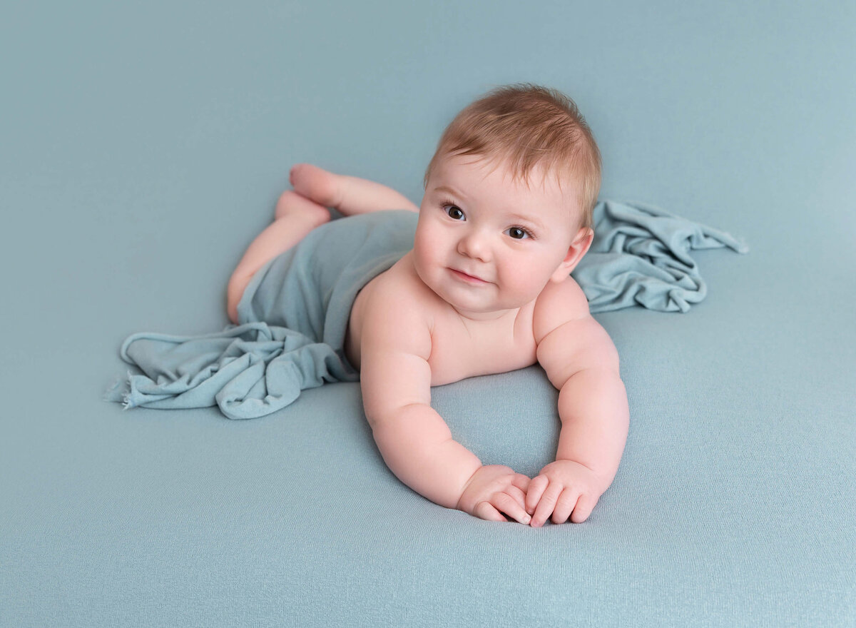 6-month-old baby boy in a teal blanket during his milestone photoshoot in NYC. Baby smiling at the camera.