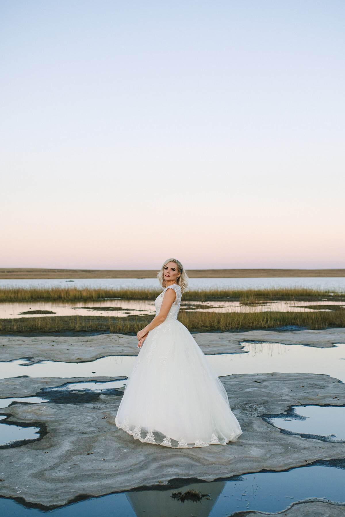 Bride standing alone on a salt pan with puddles of water, soft pink skies behind her, wearing a voluminous A-line wedding dress and looking at the camera.