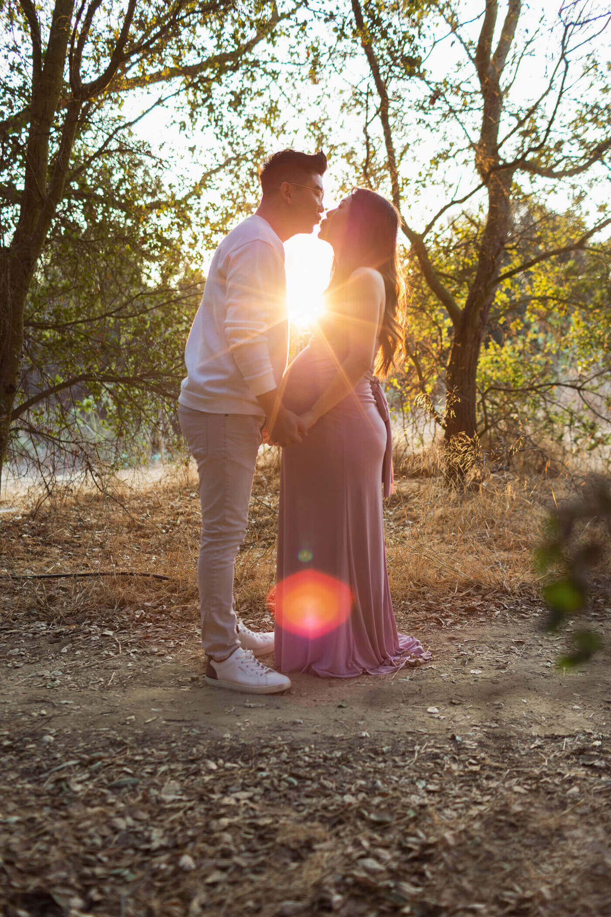 A romantic outdoor maternity portrait featuring a couple sharing a kiss at sunset. The pregnant woman, dressed in a flowing lavender gown, stands facing her partner, who wears a white sweater and light-colored pants. The sun sets behind them, casting a warm glow and creating a lens flare effect that adds a magical touch to the scene. The natural surroundings of trees and dry foliage create a serene and intimate atmosphere, highlighting the couple's love and anticipation for their baby.