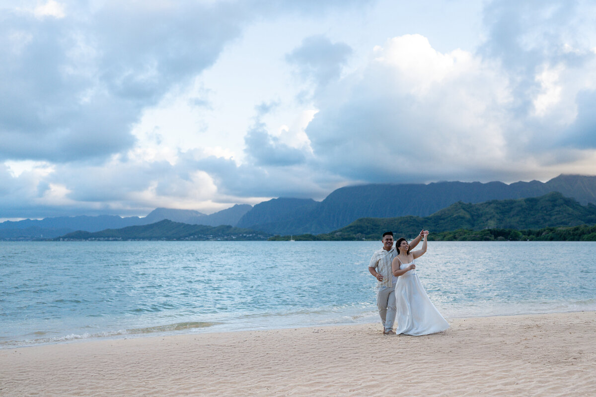Oahu Hawaii Elopement Adventurous Asian couple at Secret Island Beach during golden sunset with mountains and ocean waves dancing and spinning.