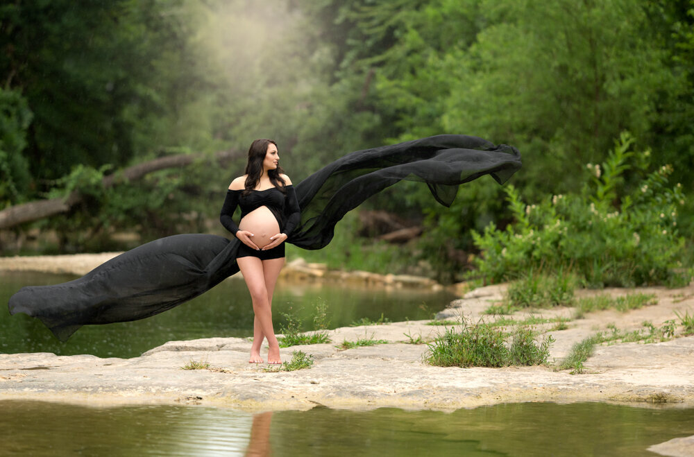 Striking maternity portrait of woman in sheer black fabric with flowing train by a creek in Austin, TX