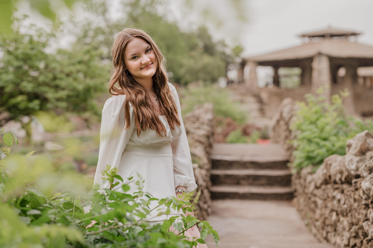 Senior picture of a girl in a white dress sitting on the edge of a stone bridge with greenery all around her.