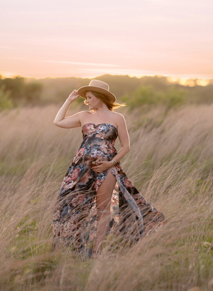Golden hour maternity photo in Cedar Park with the mom-to-be in a floral dress and hat standing in a tall grass field at sunset.