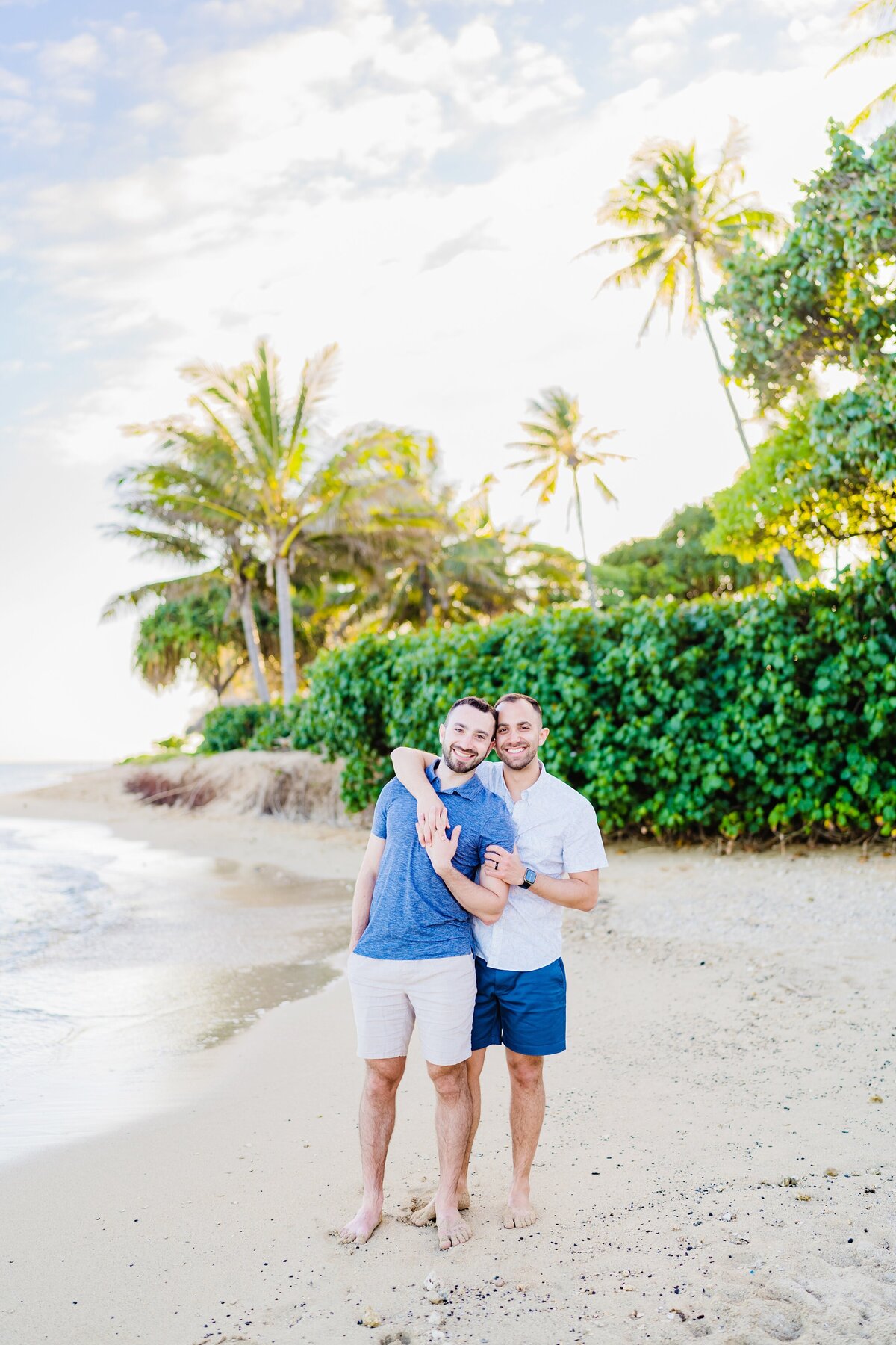 Gay couple wearing blue and white pose for a couples photo on the beach in Hawaii