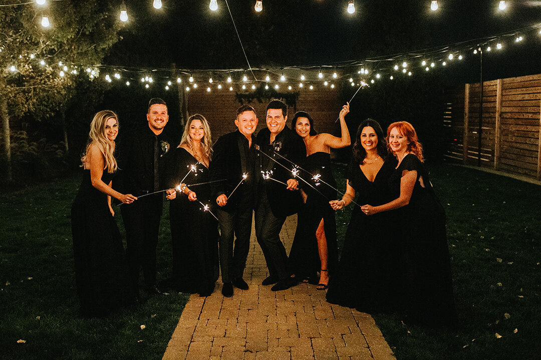 Two grooms wearing black tuxedos pose outside at night holding sparklers in hand with their wedding party.