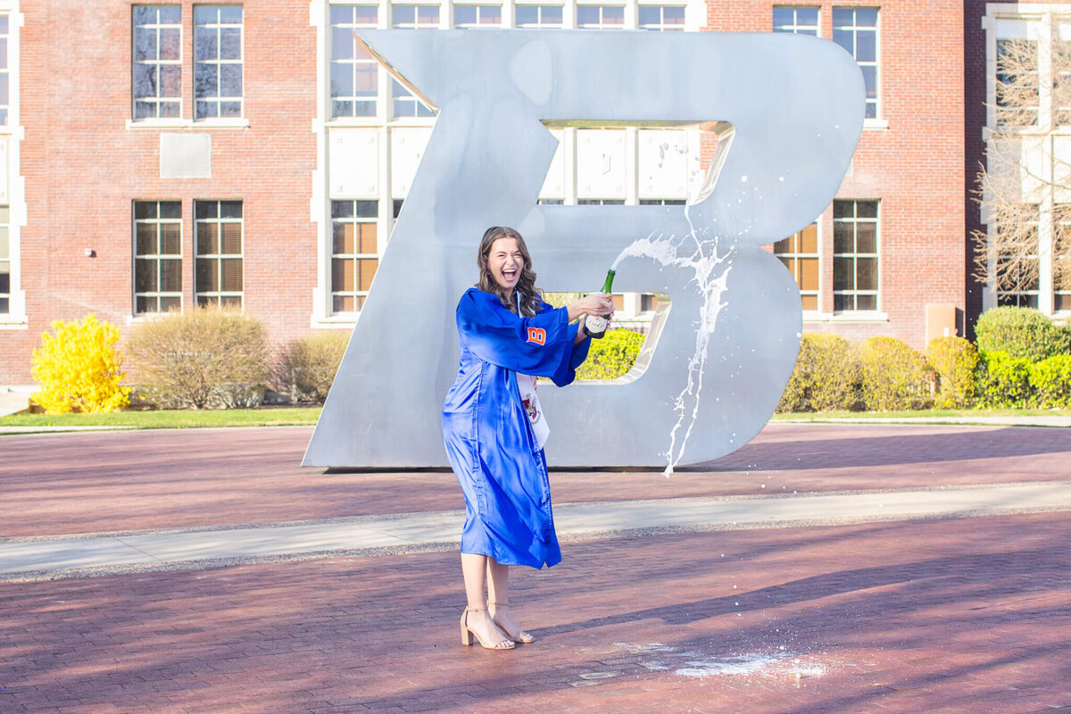 A young woman with long, brown hair wearing a dark blue graduation gown and beige heels, popping a bottle of champagne so liquid is spraying in front of her, smiling big in front of a giant "B" in front of a large campus brick building with green bushes in front of it. Taken at Boise State University.