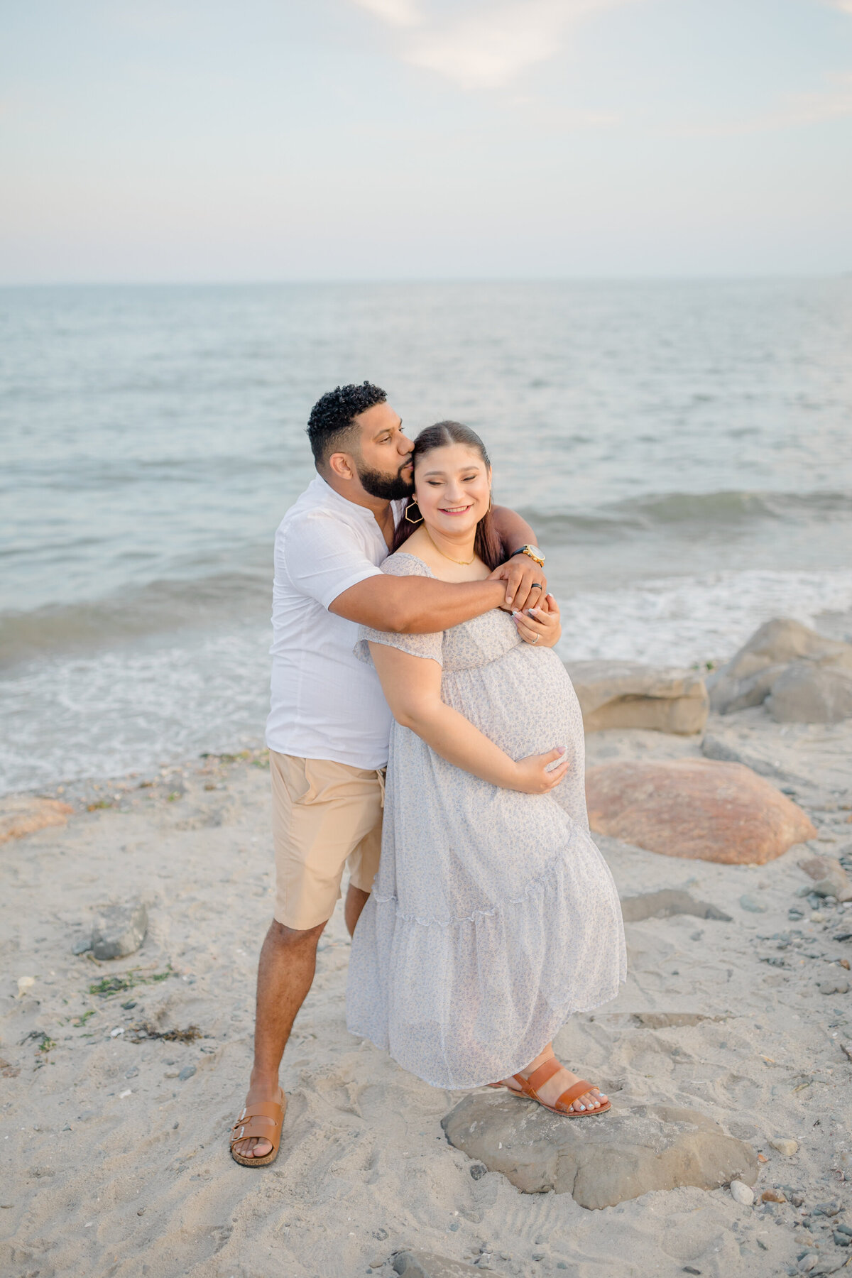 Husband and wife standing on rocks at the beach, holding hands and gazing at each other, with the ocean in the background, captured by a Connecticut maternity photographer