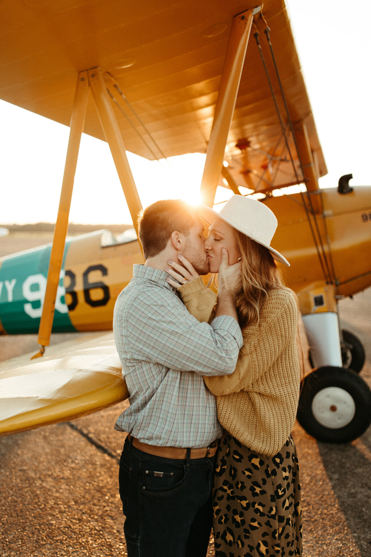 A couple is holding each other tightly and kissing as they stand in front of an airplane at an airport.