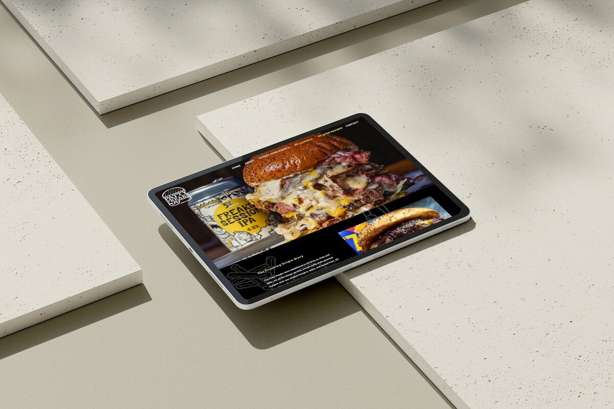 A tablet displaying the Patty Freaks website, showcasing a close-up image of a juicy, overflowing burger with melted cheese and bacon, alongside a can of "Freak Session IPA". The screen highlights their menu and food offerings. The tablet is placed on a light-coloured, textured surface with geometric shadows, creating a stylish and appetising presentation.