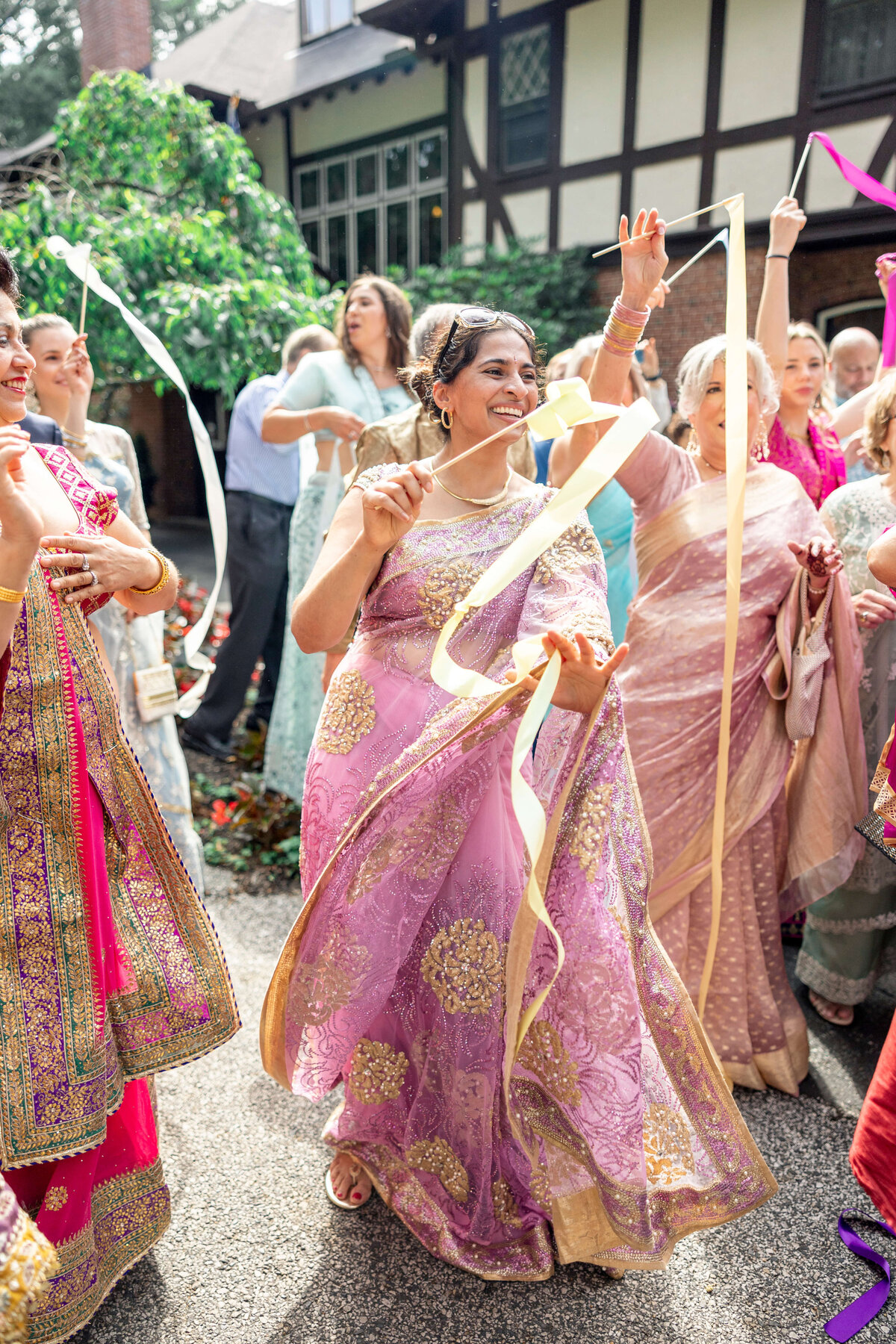 A group of people in colorful traditional attire joyfully dance with ribbons in an outdoor setting. The central figure wears a purple and gold sari, smiling and engaged in the celebration. Tudor-style building visible in the background.