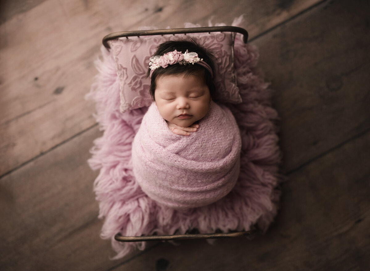 Baby girl is wrapped in a pink knit swaddle and plated atop of a miniature newborn prop bed for her Riverside, CA newborn photoshoot. Baby is sleeping an her fingers are peeking out of the swaddle. Captured by best Riverside newborn photographer Bonny Lynn Photography.