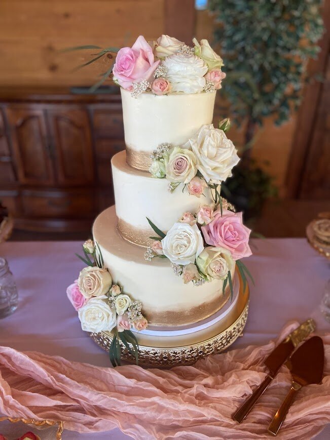 A three tier cake is shown on a white tablecloth. The cake is white with a painted gold band around the bottom of each tier. There's clusters of white and pink fresh flowers.