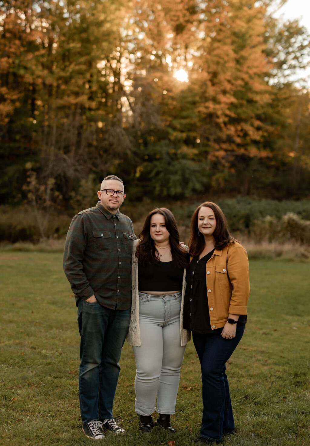 Three people stand together on the grass with autumn trees in the background, captured perfectly by a talented New England portrait photographer. The sun peeks through the trees as they pose casually in jeans and tops, two wearing jackets, embodying the essence of portrait photography in New England.