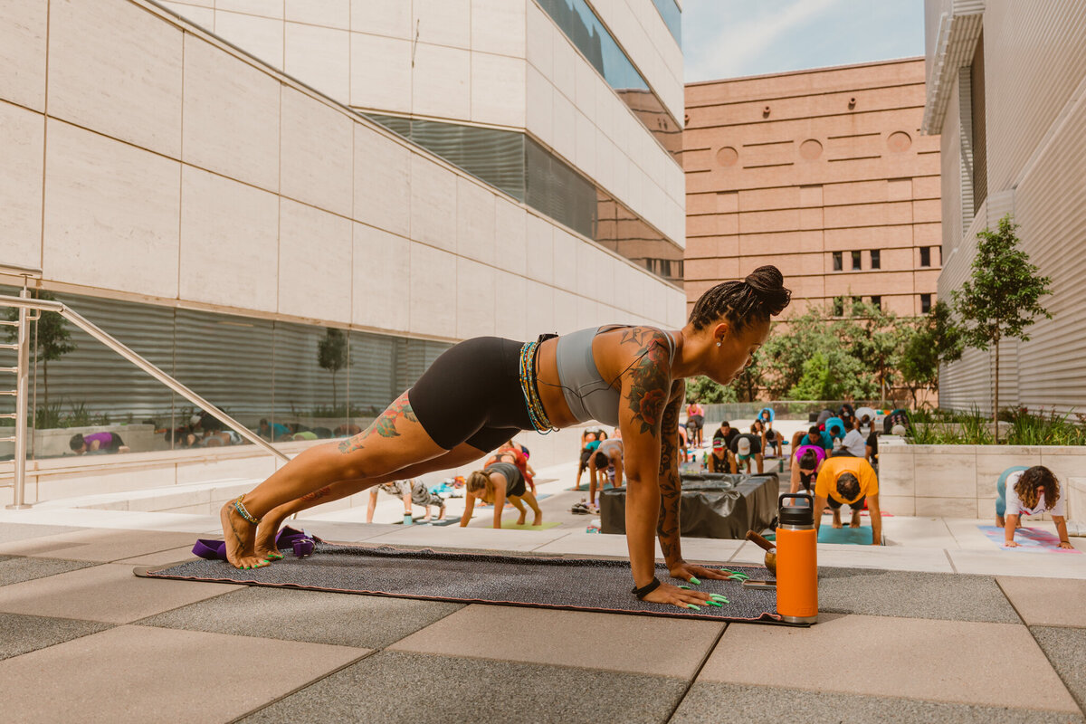 Woman holding a plank pose during the Yoga and Mimosas outdoor event in Houston, Texas, with a group of participants in the background. 