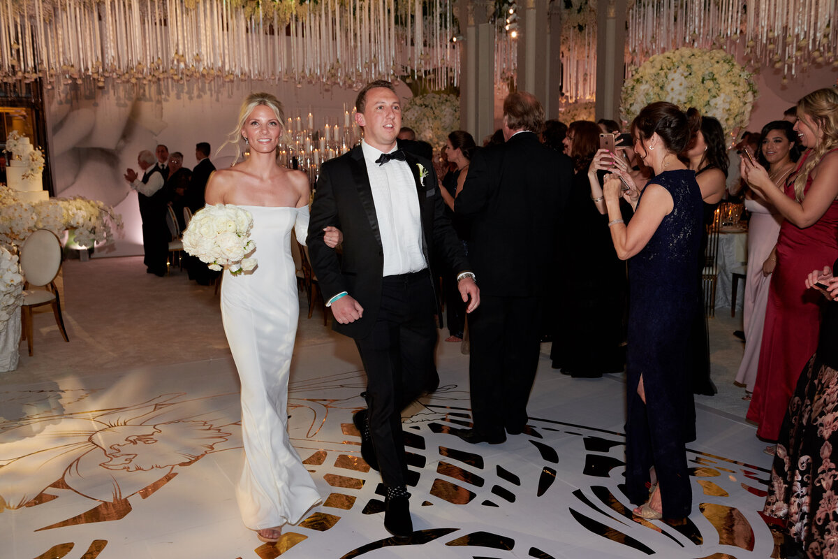 The photo was taken at the iconic New York City landmark, The New York Public Library, by the talented photographer Christian Oth. The beautiful bride and groom, both beaming with happiness, stroll hand in hand at their wedding reception. The bride looks stunning in a white off-the-shoulder gown, delicately holding a bouquet of white flowers. Surrounding them are guests dressed in formal attire, enthusiastically applauding and capturing the special moment with their cameras. The venue itself is adorned with elegant decorations, adding to the atmosphere of joy and celebration. Videographers from East West Films were also present to capture the magical moments of the day.