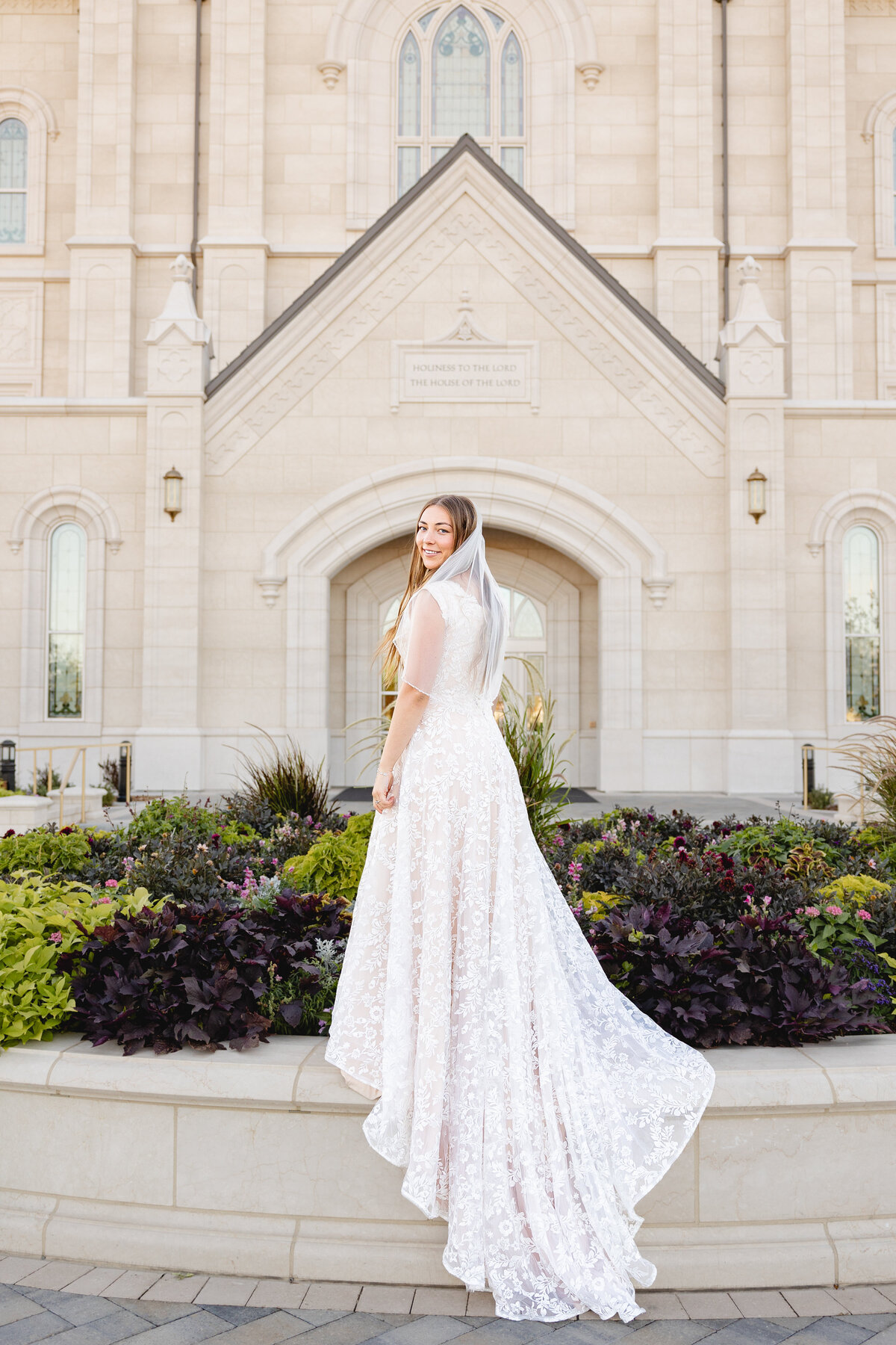A bride in a lace wedding dress and veil smiles while standing in front of the Taylorsville Utah LDS Temple, surrounded by colorful flowers and greenery.