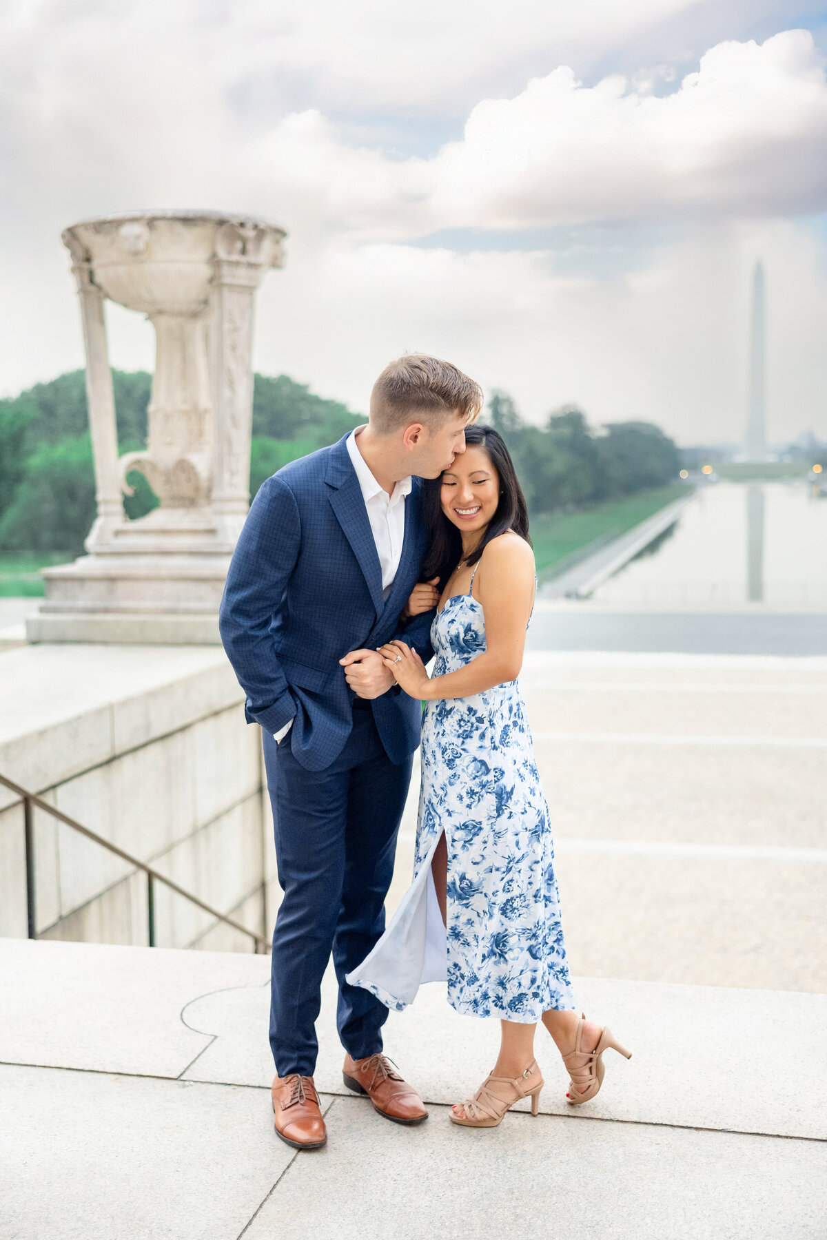 A couple stands on a terrace near the Washington Monument. The man, in a blue suit, gently kisses the womans forehead. She wears a blue floral dress and smiles, with the reflecting pool visible in the background on a cloudy day.
