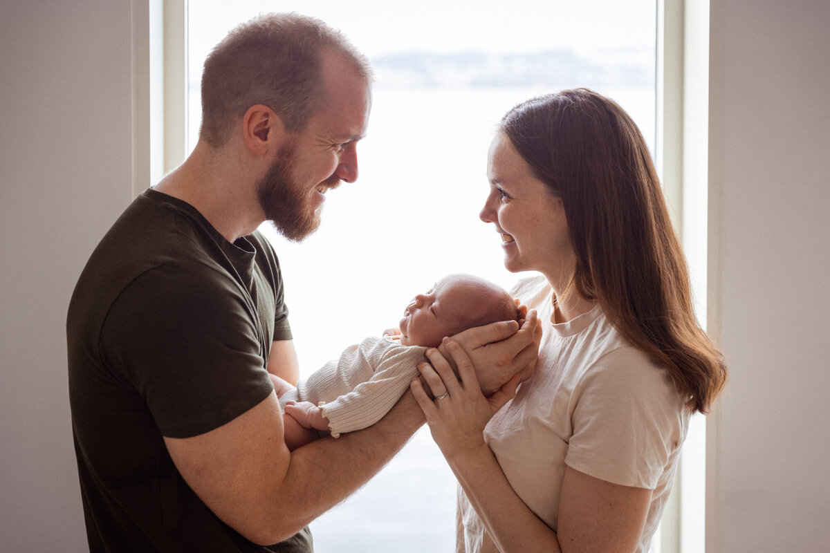 Livsstilsfoto familieportrett tatt ved vindu. Far og mor står ved vindu, mor holder baby, far stryker baby på hodet.