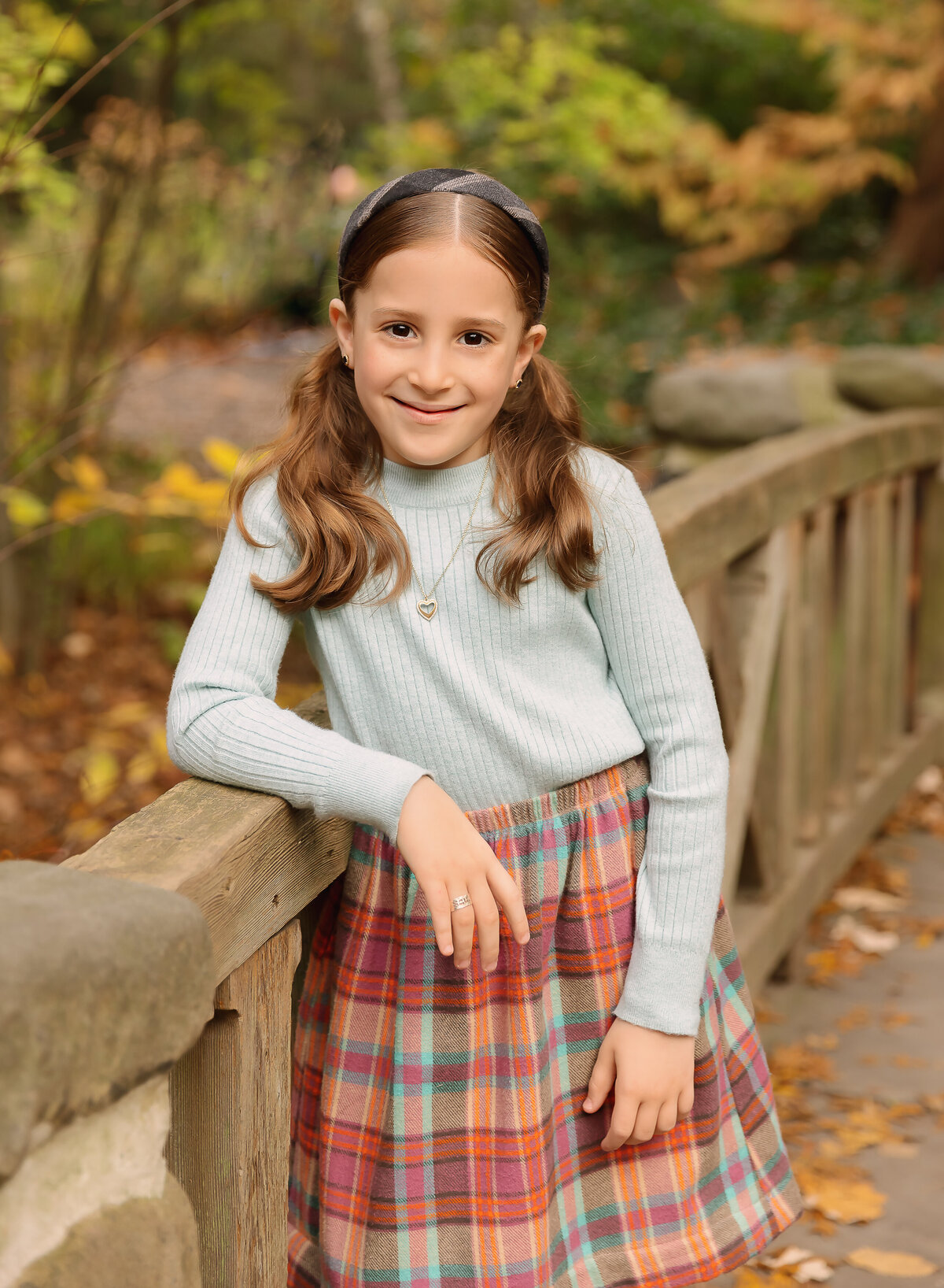 Little girl in grey sweater and plaid skirt is stanging on a stone bride for a fall family photoshoot. Her arm is resting atop of the bridge and she is smiling at the camera. Captured by Brooklyn family photographer Chaya Bornstein