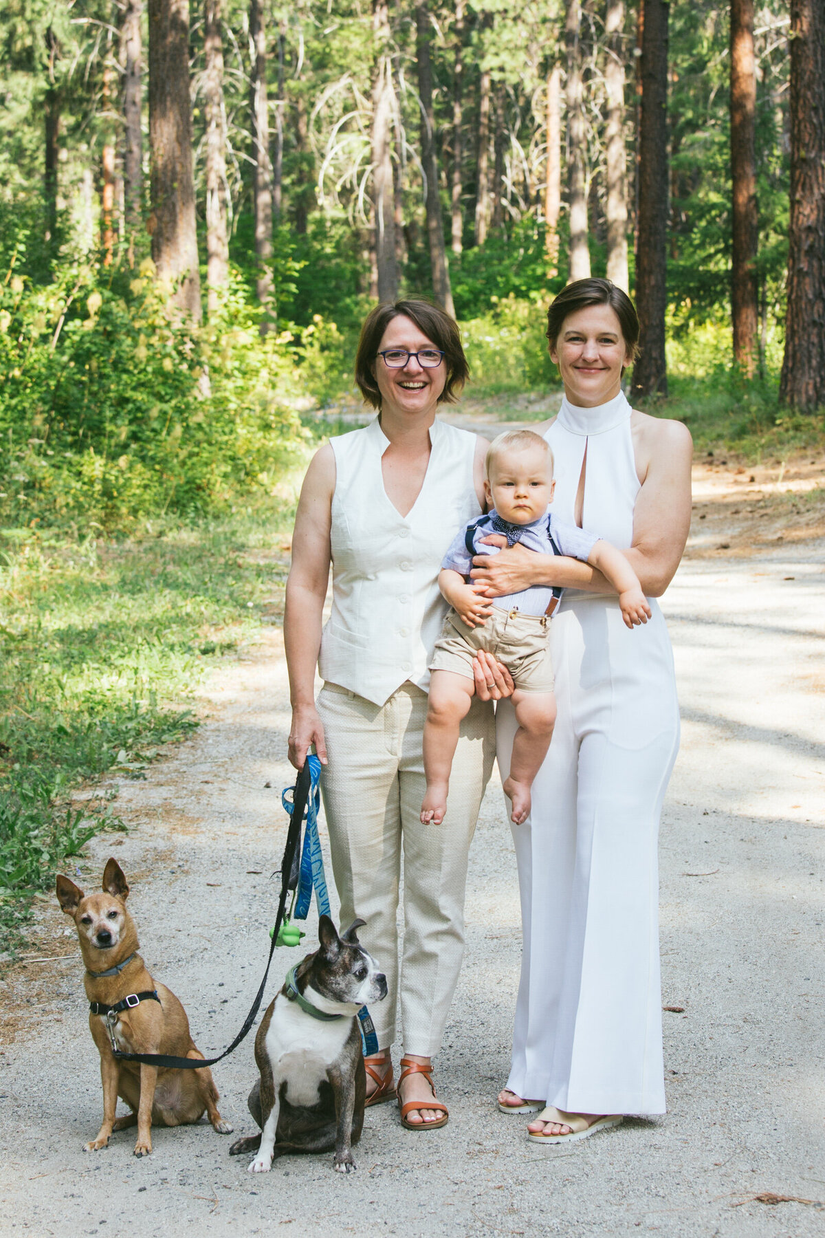 Two brides standing on a trail, holding their son and two dogs for a wedding photo.
