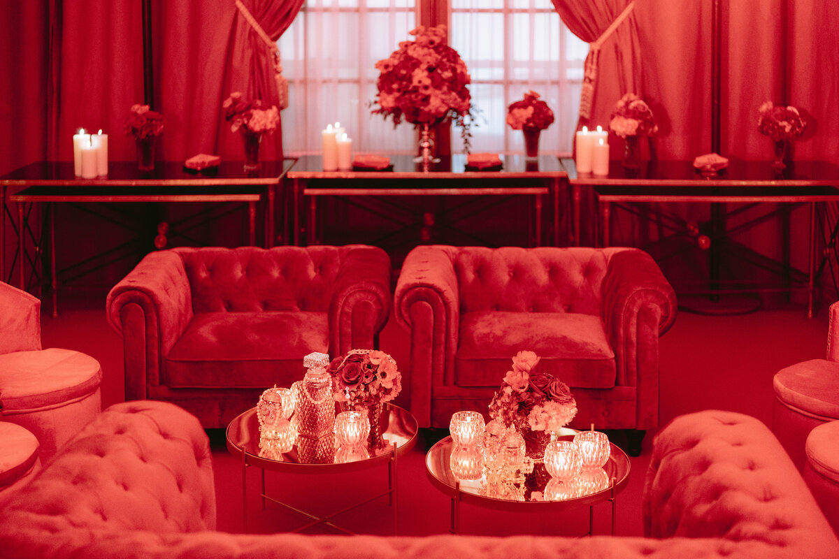 The photo showcasing a room bathed in red light at the luxurious Shangri La Hotel & Ritz Paris Hotel in Paris, France was taken by the talented photographer Matteo Coltro. Videographers Zen Film Works also captured stunning footage of the elegant setup, highlighting the intricate details and ambiance of the scene. In the image, plush velvet chairs are elegantly arranged around circular glass tables, each adorned with exquisite flower arrangements and softly lit candles. The backdrop is enhanced by additional tables displaying more beautiful floral decorations and flickering candles, while striking red curtains elegantly frame a grand window, adding a touch of drama and sophistication to the overall scene.