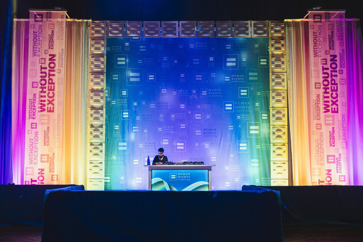 A DJ plays music in front of a rainbow technicolor background.