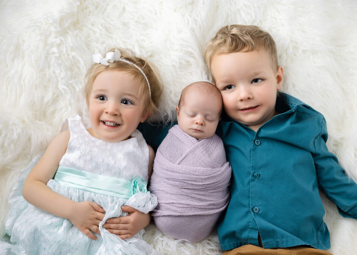 young brother and sister laying on a white shaggy rug with their new baby sister wrapped in purple fabric