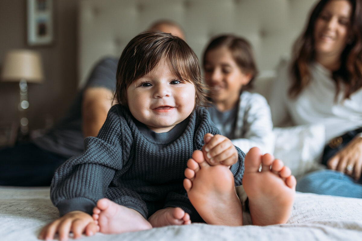 Family Photographer, on a bed, a little smiling baby grabs his sisters toes, mom and dad behind them