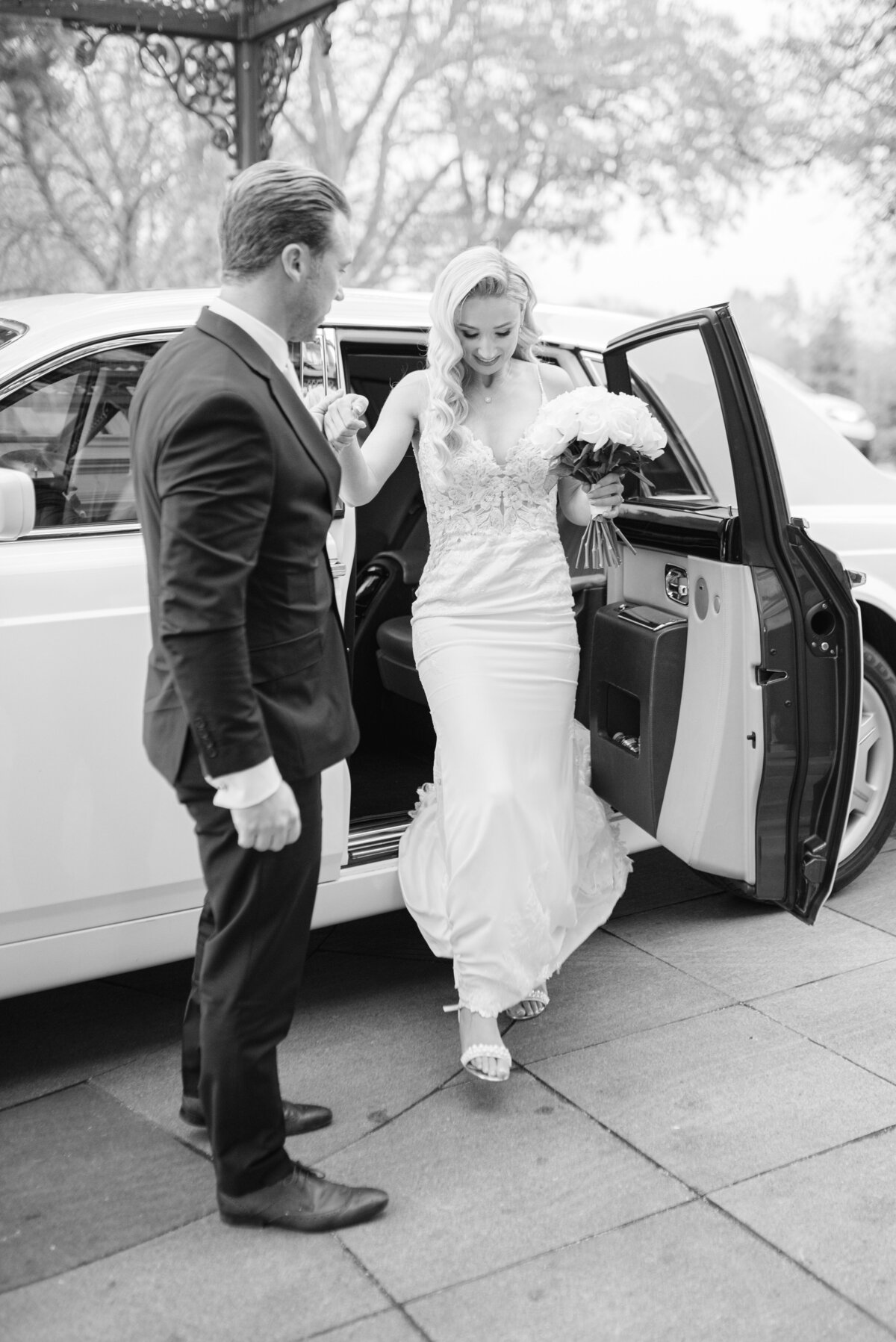 The groom is helping the bride get out the wedding car at the hotel. the image is edited in black and white
