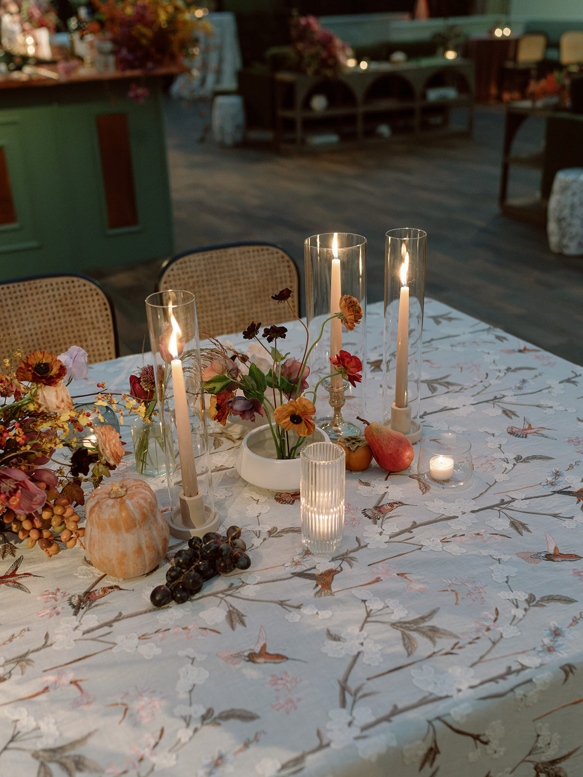 Clustered and styled floral arrangements, fruits, and art pieces for fall wedding reception. Low centerpieces mixed with ikebana arrangements and bud vases decorate the reception tables at this fall wedding. Floral centerpieces featuring orchids, ranunculus, sweet peas, and bittersweet. Styled wedding tables with fruits, curated candles, and floral arrangements. Tasteful fall color palette of copper, ruby, gold, burgundy, rust, and lavender. Design by Rosemary & Finch Floral Design in Nashville, TN.