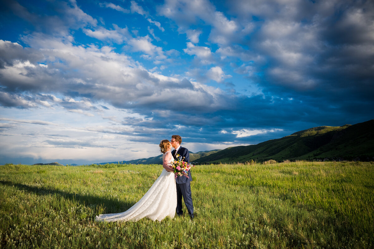 A wide angle shot of a bride and groom kissing in a grassy field with a Colorado blue sky in the background.