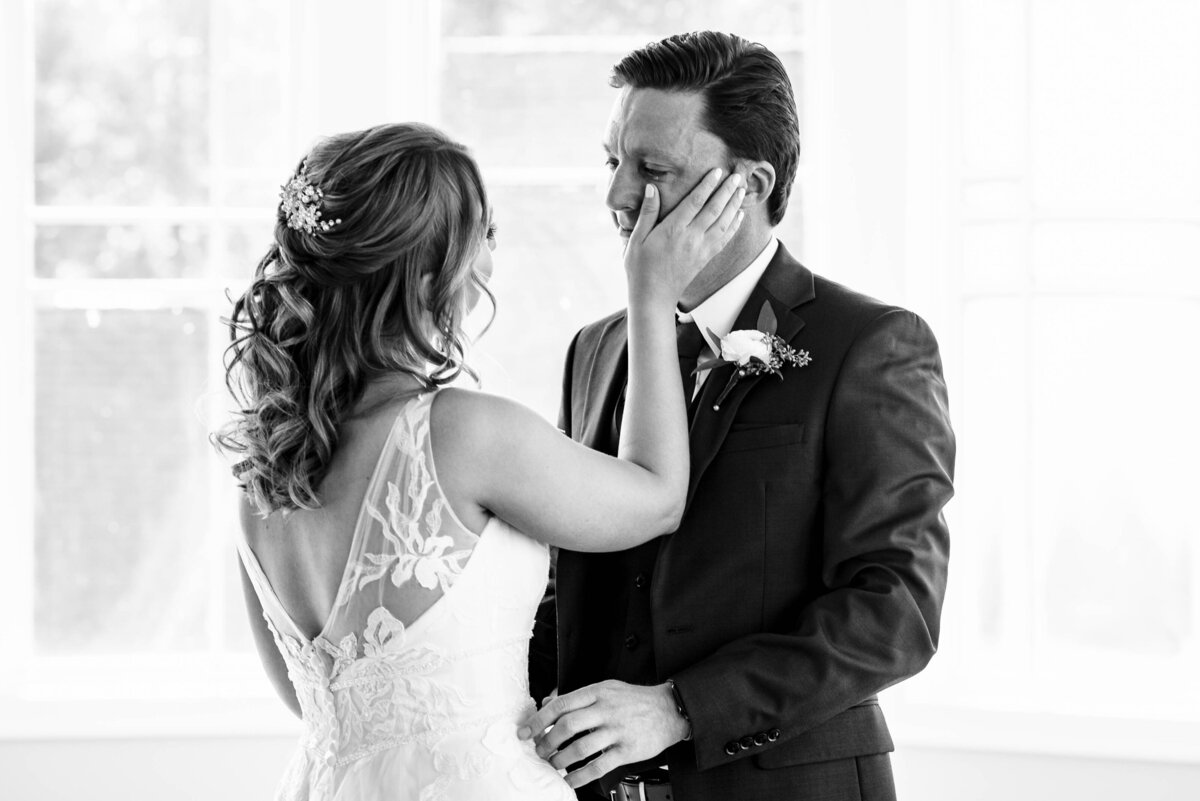 Black and white image of bride wiping tear from groom's face during first look at Pine Island Country Club by Charlotte wedding photographers DeLong Photography