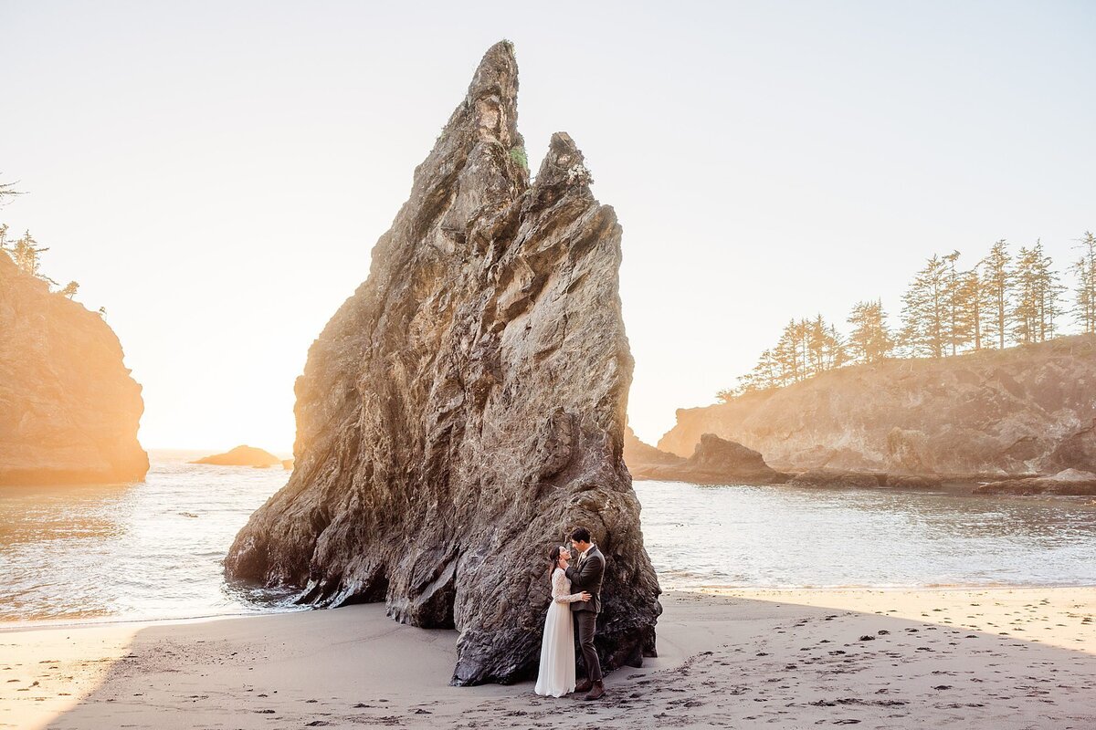 A bride and groom stand next to a giant sea stack on the beach.