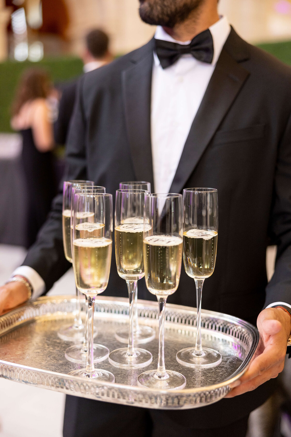 A waiter in a suit and bow tie holds a silver tray with several filled champagne glasses. The background is slightly blurred, suggesting a formal event or celebration.