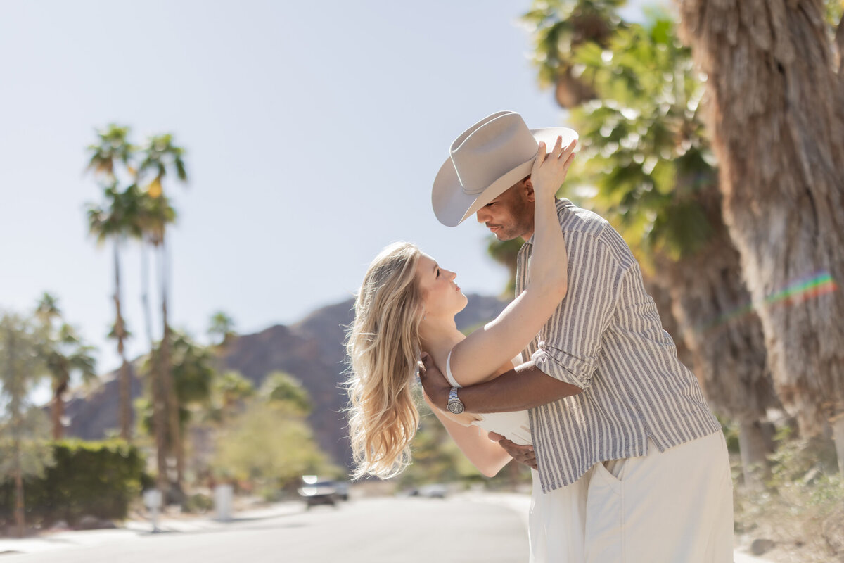 Joyful couple twirling in a sunny Palm Springs setting, dressed in neutral tones with mountains in the distance.