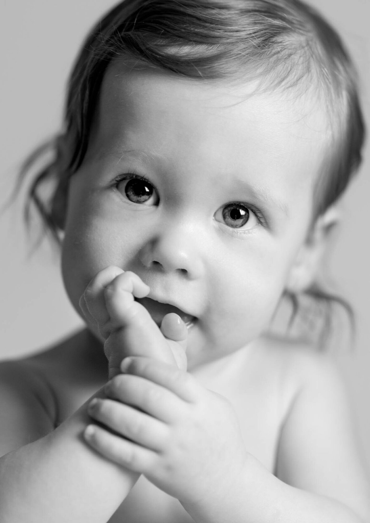 A black-and-white photo of a curious baby holding their foot with both hands. The baby's large eyes and slight smile convey innocence and wonder.