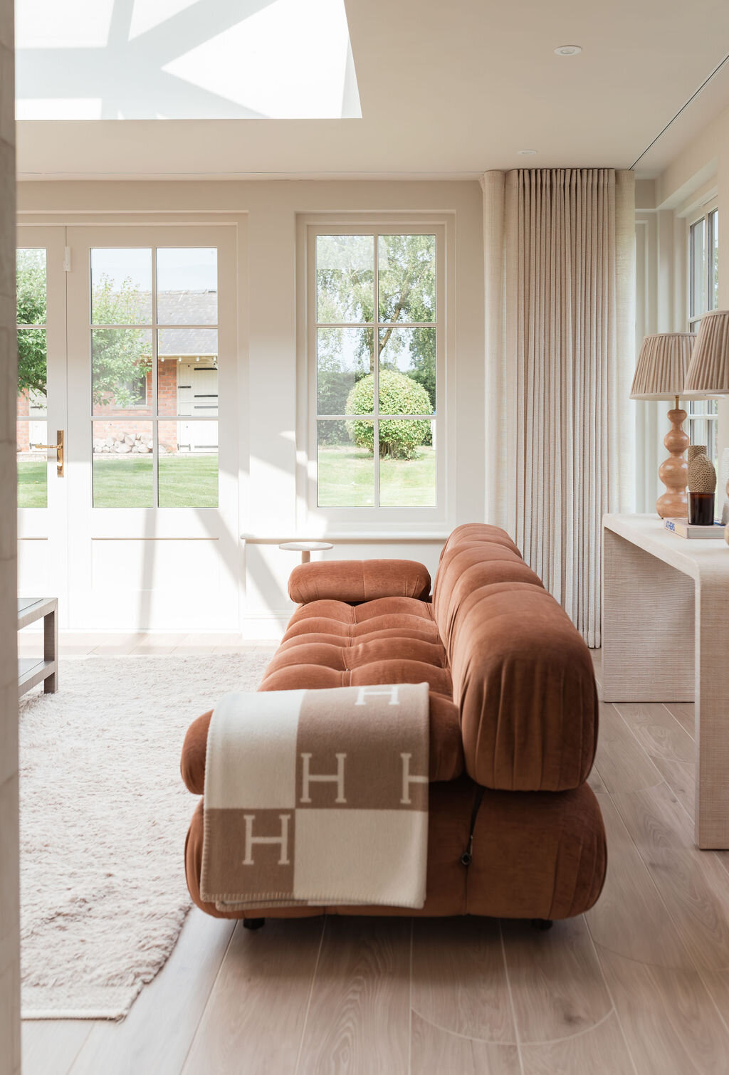 A cozy living room with large windows features a plush, rust orange tufted sofa, a light-colored rug, and a wooden coffee table adorned with decorative items and books. Two large, neutral lamps sit on a console table behind the sofa, and greenery is visible outside.