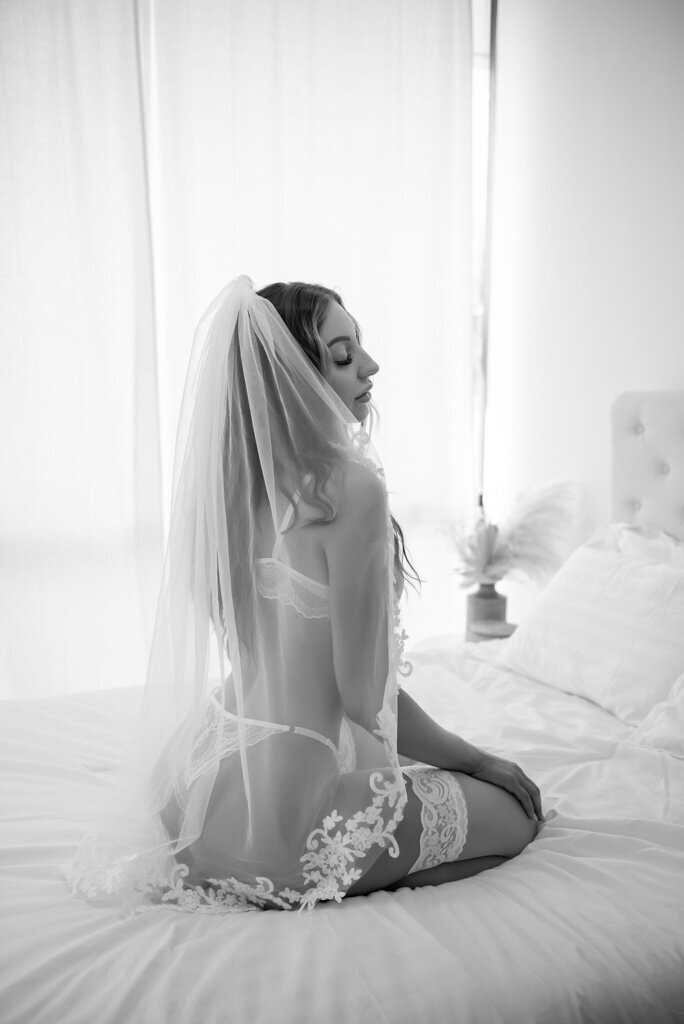 Black and white portrait of bride wearing lingerie and a veil kneeling on a white bed looking over her shoulder