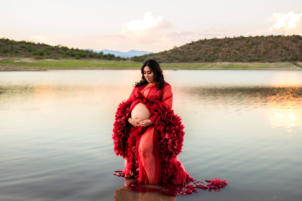 Beautiful pregnant mom in red couture gown standing in water at lake. Captured by Cactus & Pine Photography 